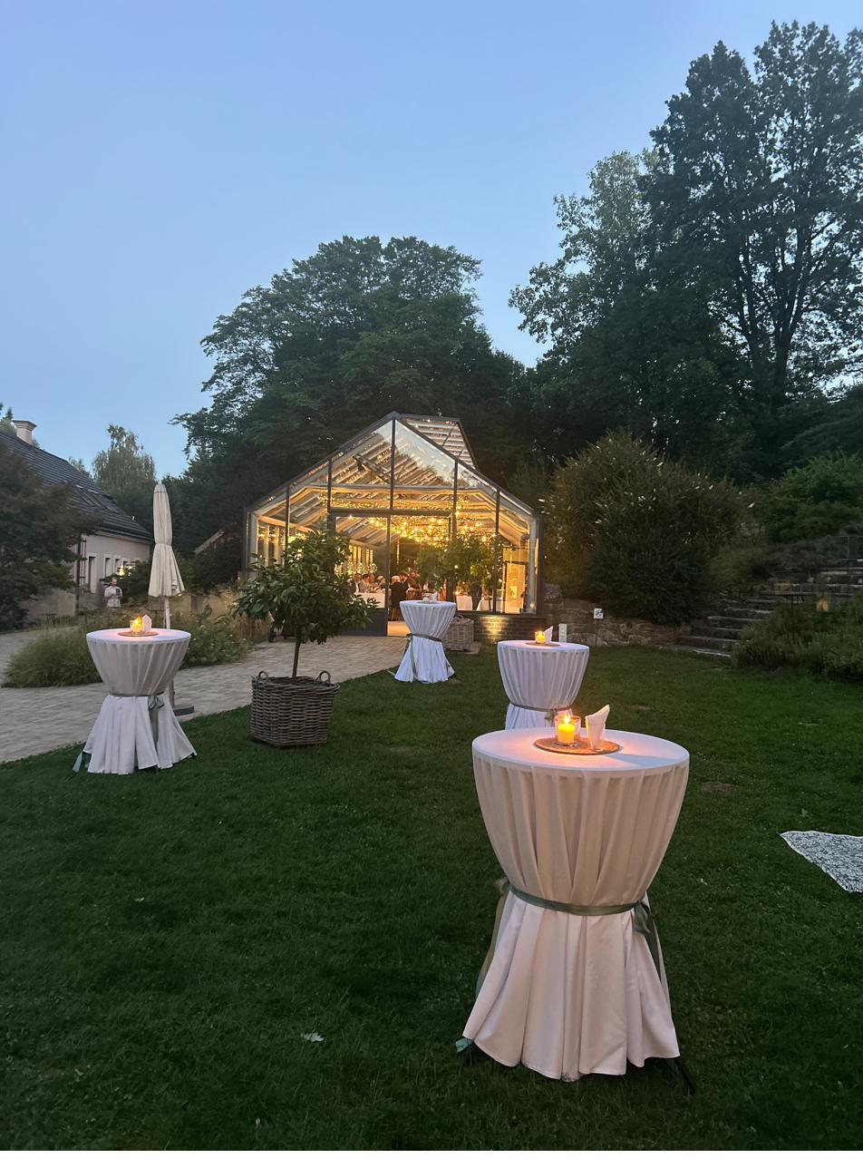Beleuchtetes Glashaus in der Abenddämmerung mit Stehtischen im Garten
