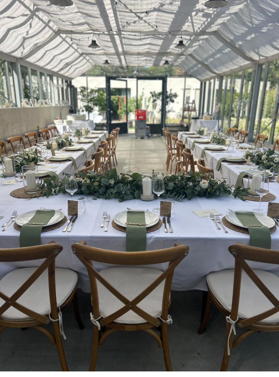 Lange Hochzeitstafel in einem Glashaus, gedeckt mit weißem Leinen, Eukalyptus und Holzstühlen